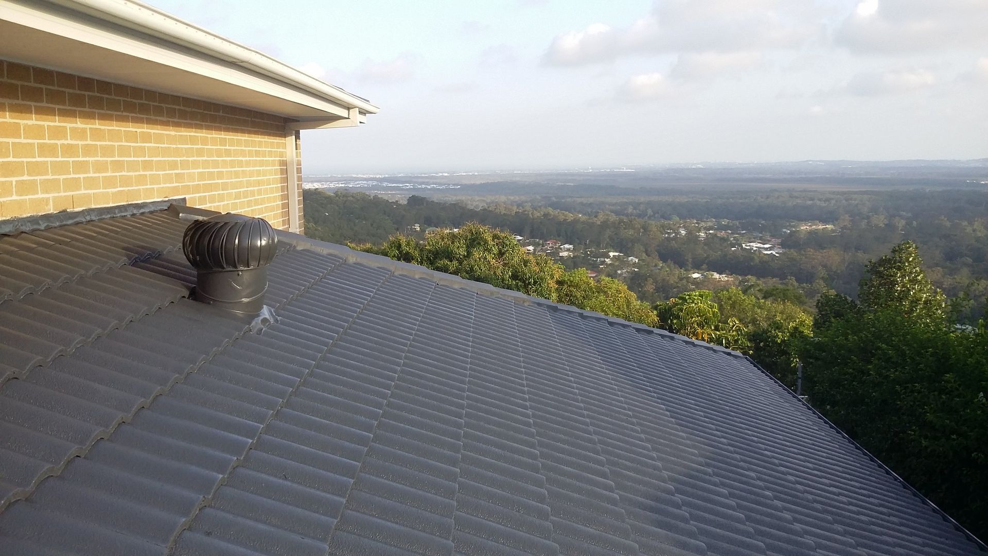 Dark gray tiled roof with a rotating vent, overlooking a green landscape with a distant cityscape.