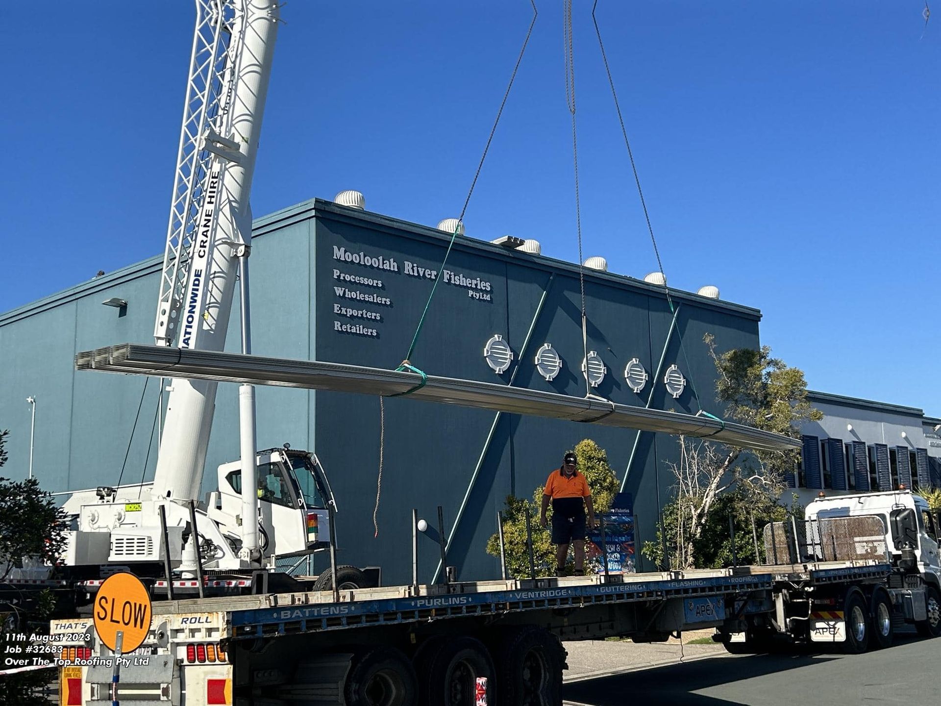 A crane lifts a large, horizontal structural beam from the back of a flatbed truck near a blue industrial building.