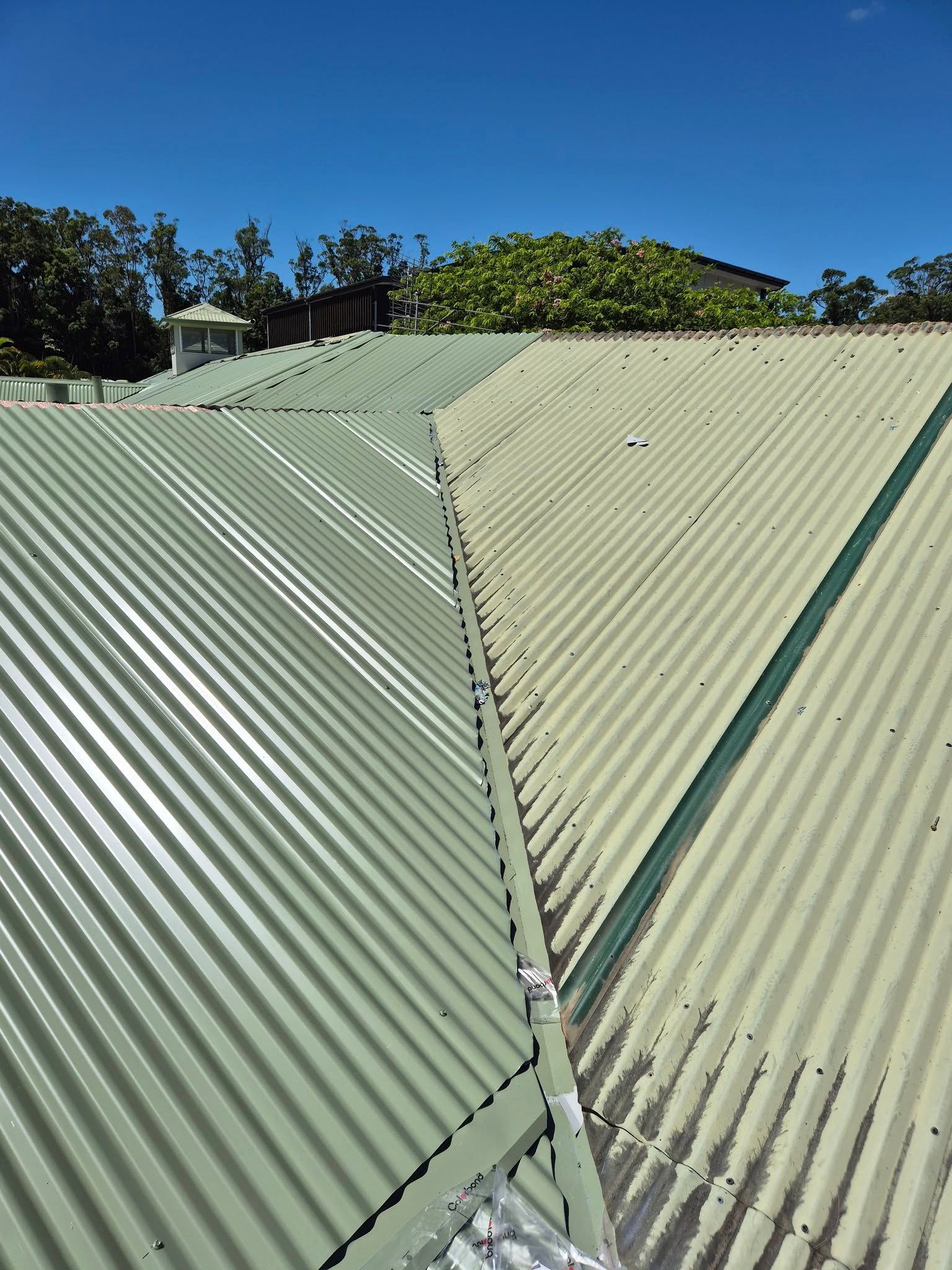 Green Metal Roof Under a Blue Sky, With Trees Visible in the Background — Over the Top Roofing Specialists in Narangba, QLD