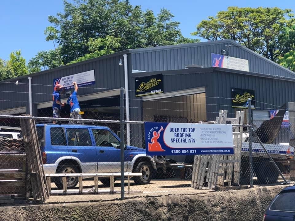 Two People Installing a Sign on a Building. Blue SUV Parked in Front — Over the Top Roofing Specialists in Nambour, QLD