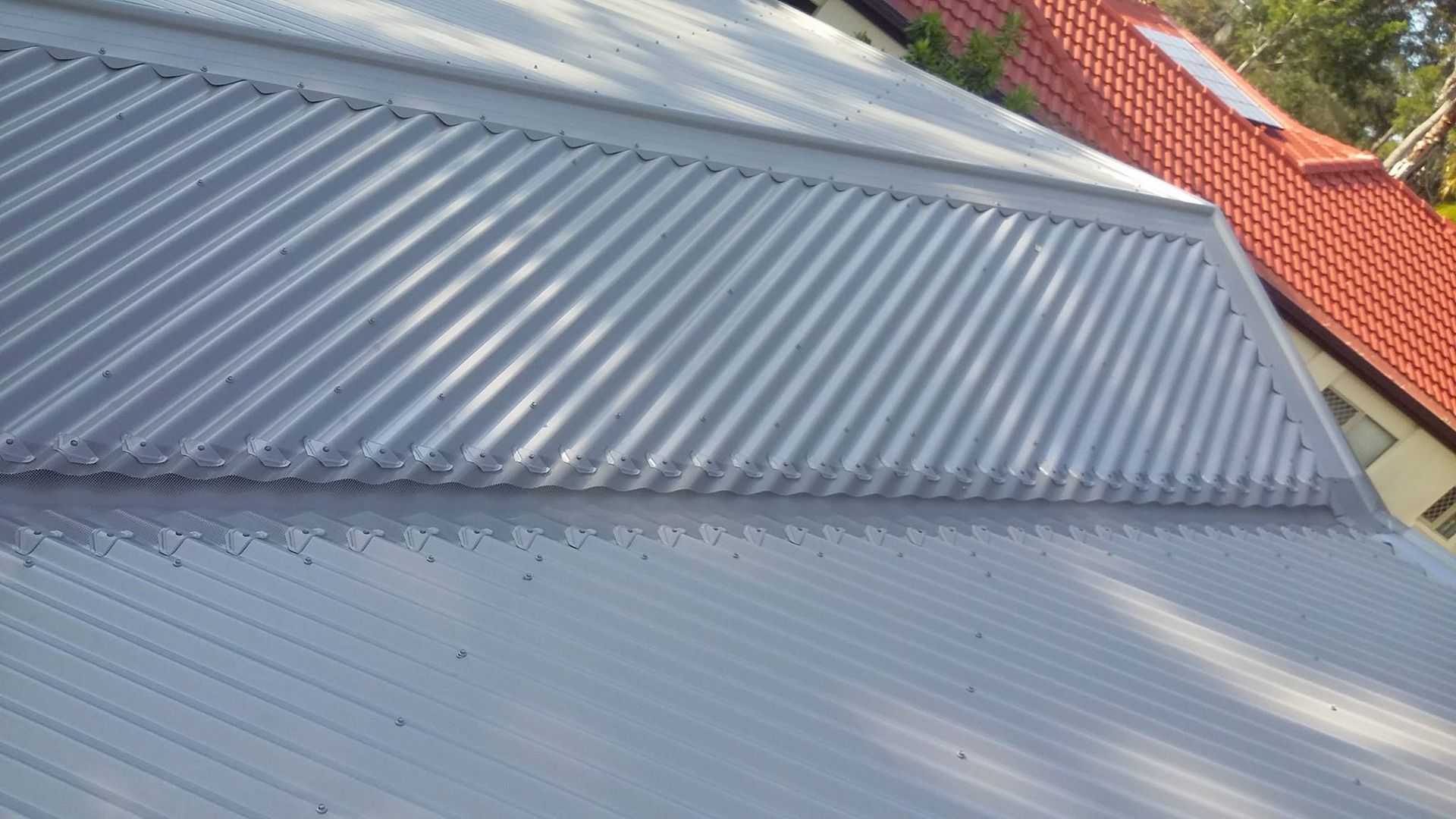 Gray Metal Roof With Diagonal Ridge and Red-tiled Roof in Background — Over the Top Roofing Specialists in Buderim, QLD