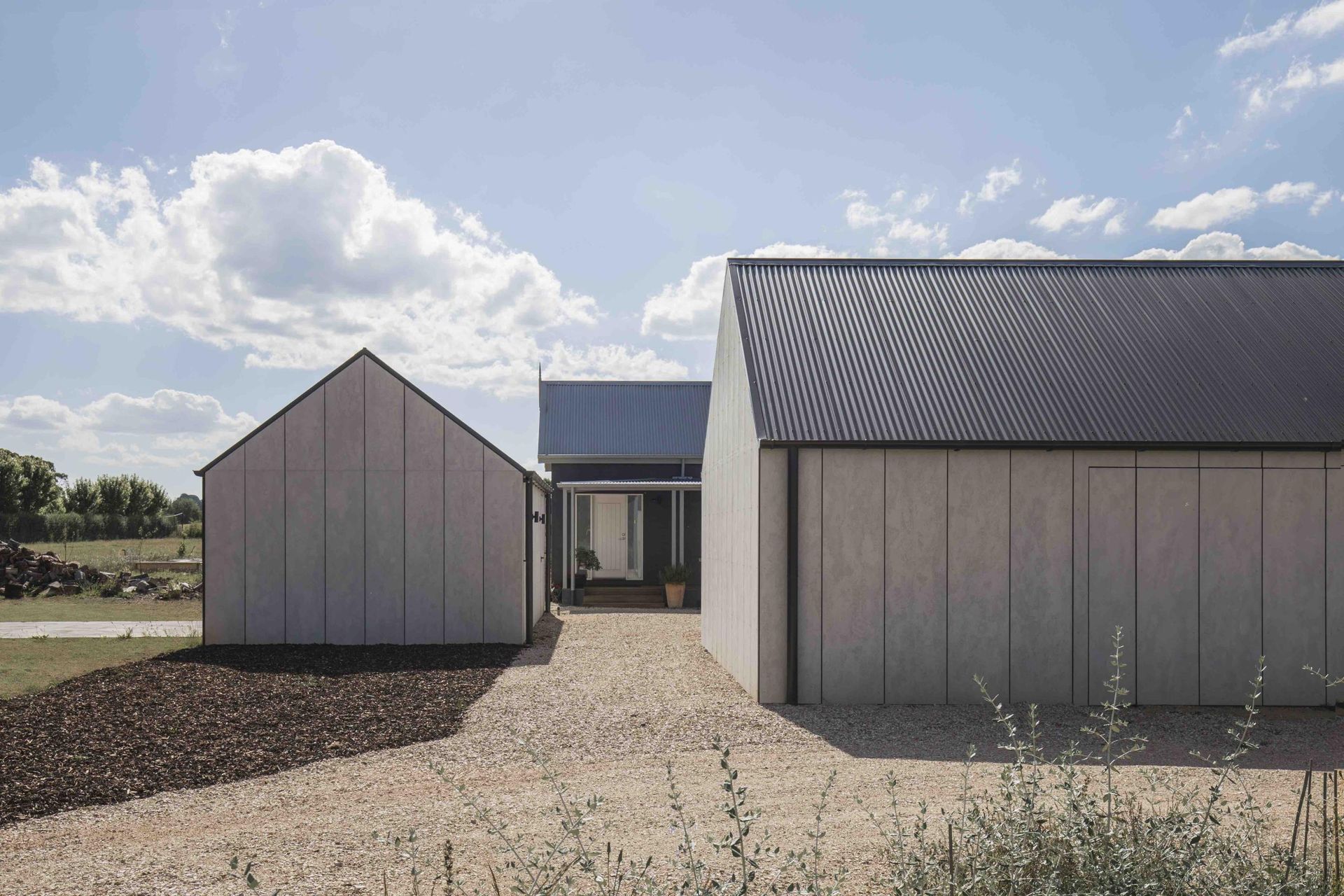 Three modern, light-gray structures with dark roofs. Gravel pathway leading towards central open area under a blue sky.