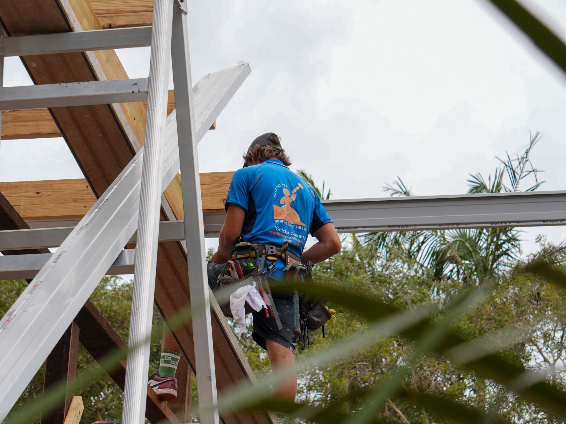 Construction worker climbing a ladder to a roof, wearing a blue shirt and tool belt.