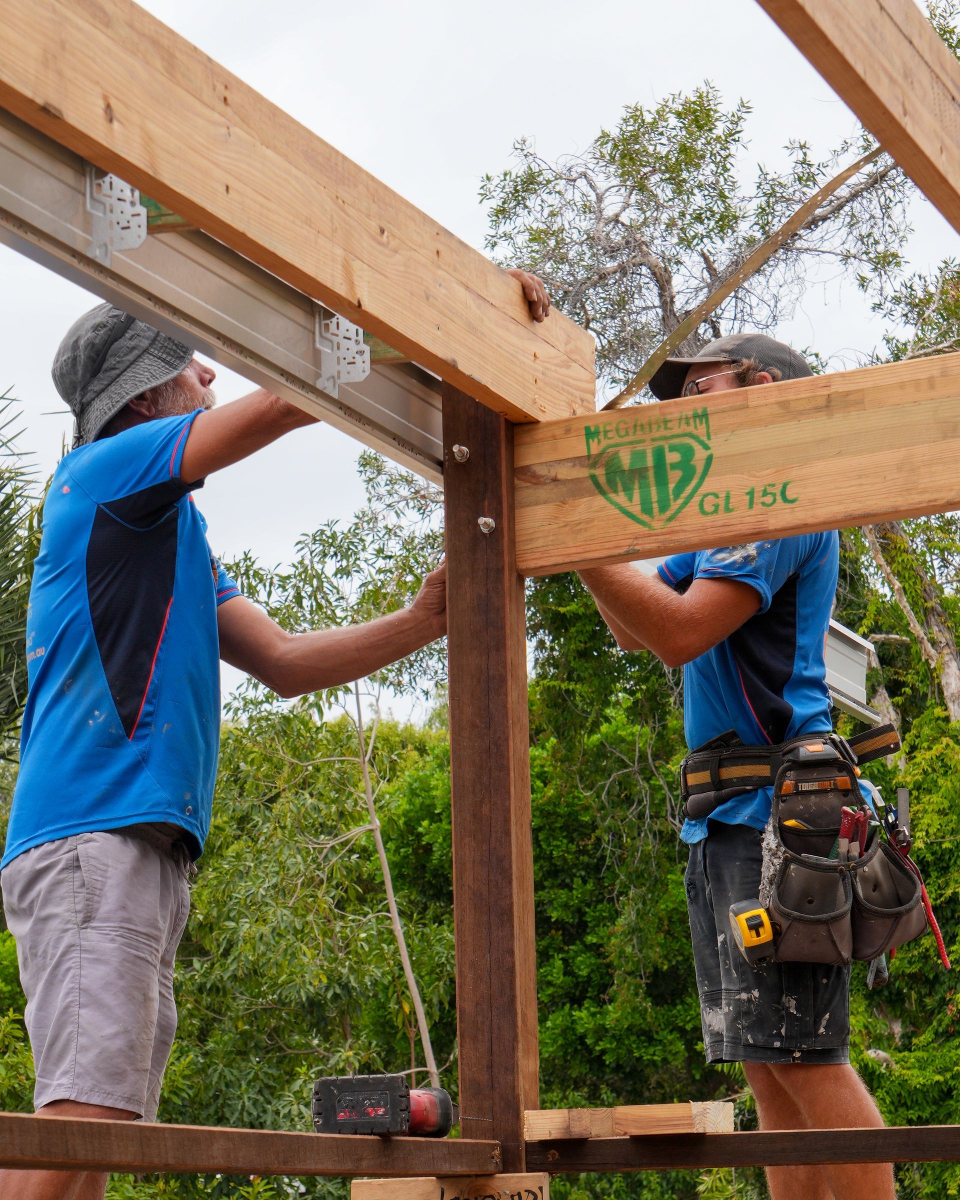 Two people building a wooden structure outdoors. They are connecting beams. One holds a beam in place.