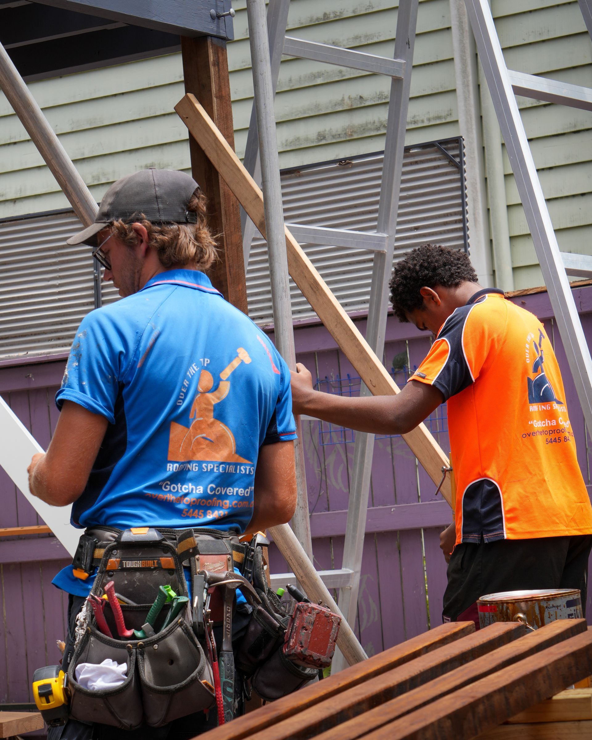 Two construction workers building, one in blue, the other in orange, working near a wooden fence.