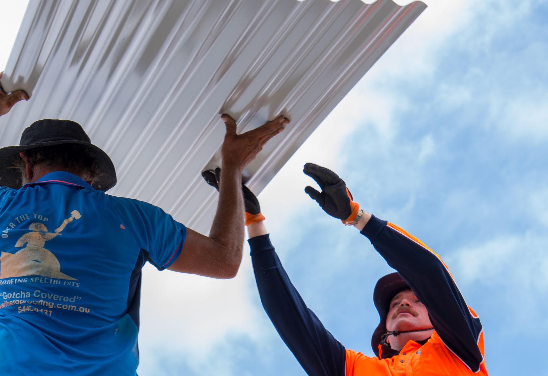 Two construction workers in high-visibility gear lift a corrugated metal sheet against a blue sky.