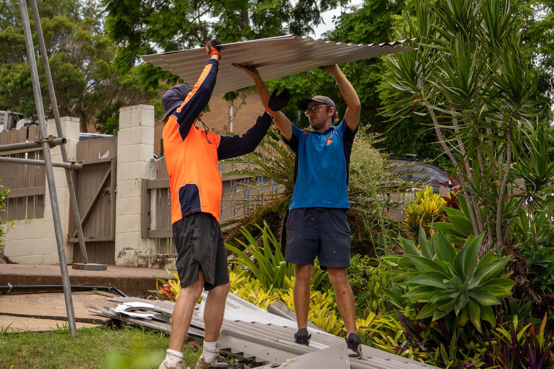 Two people holding up a corrugated metal sheet outdoors, near a building.