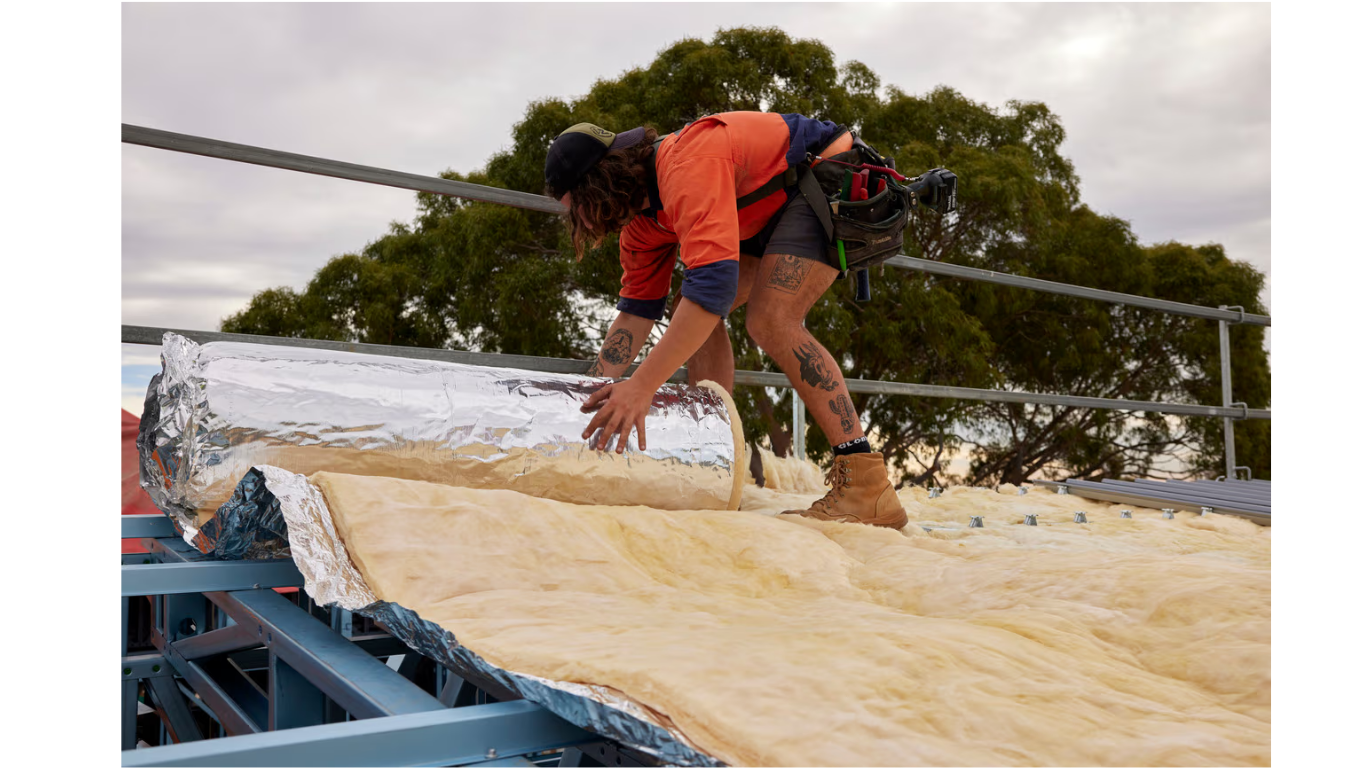 A person in high-visibility workwear installs reflective insulation batts onto a metal roof frame.