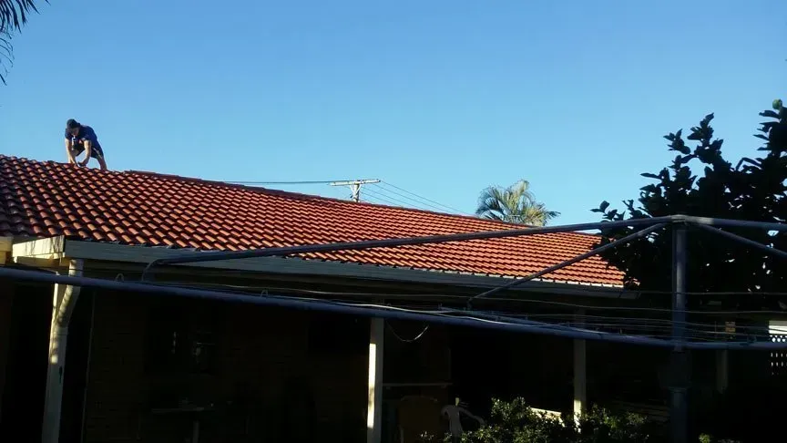 Person on a Tiled Roof Under a Blue Sky, Near a Clothesline and House — Over the Top Roofing Specialists in Narangba, QLD