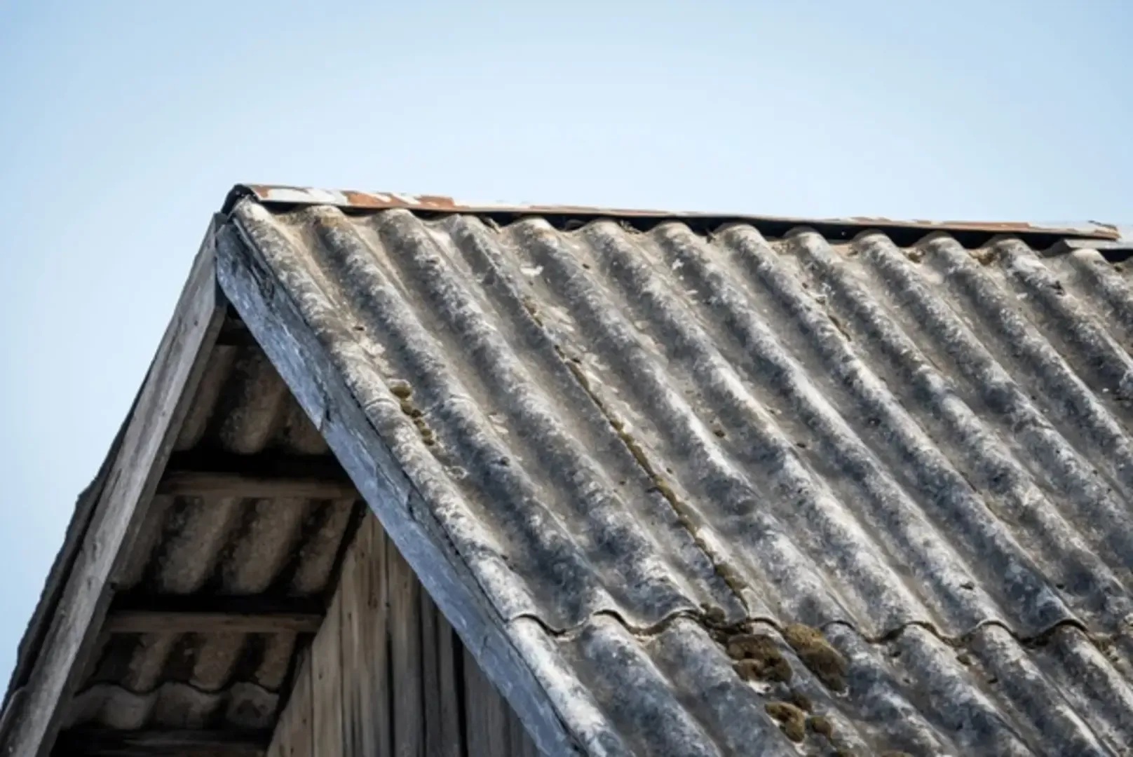 A close-up view of an old, weathered, gray corrugated fiber-cement roof with moss, set against a clear blue sky.