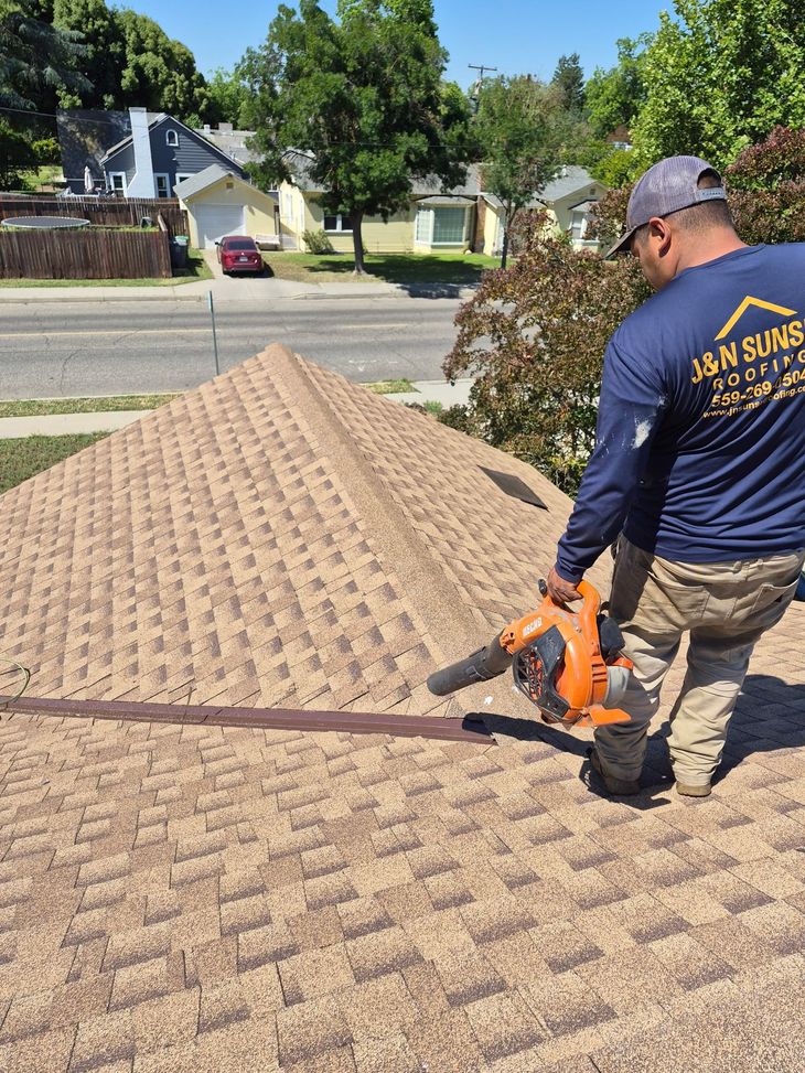 Worker using an orange leaf blower on a shingled roof in a residential neighborhood