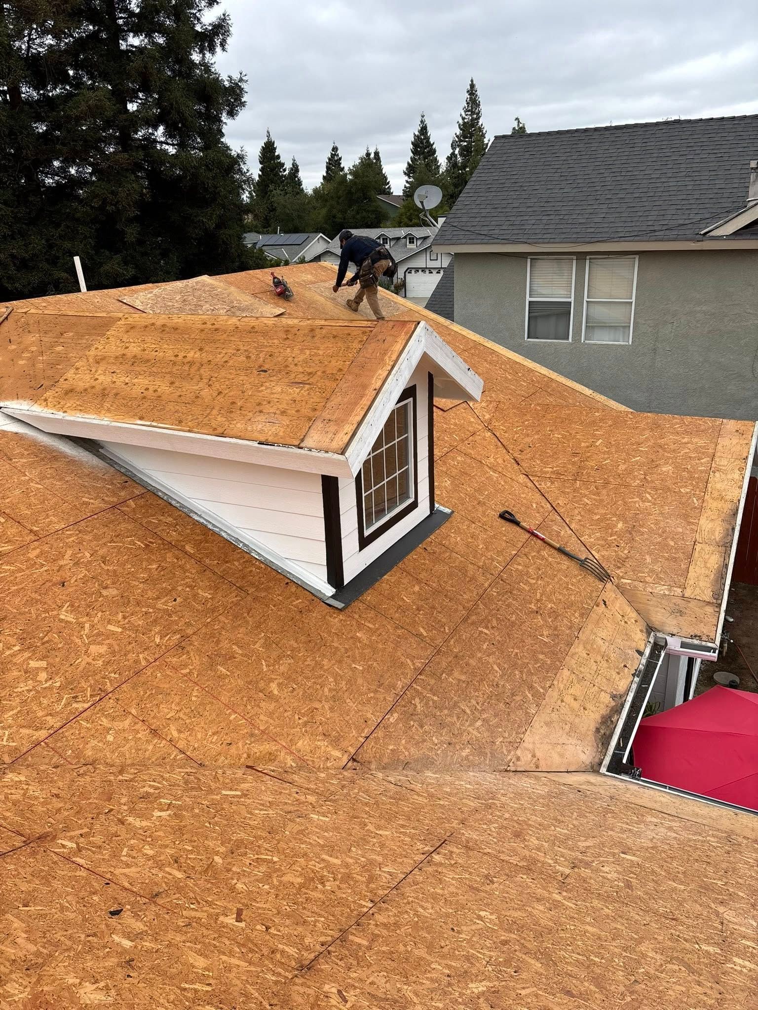 Workers on a roof installing brown roofing underlayment beside a dormer in a suburban neighborhood