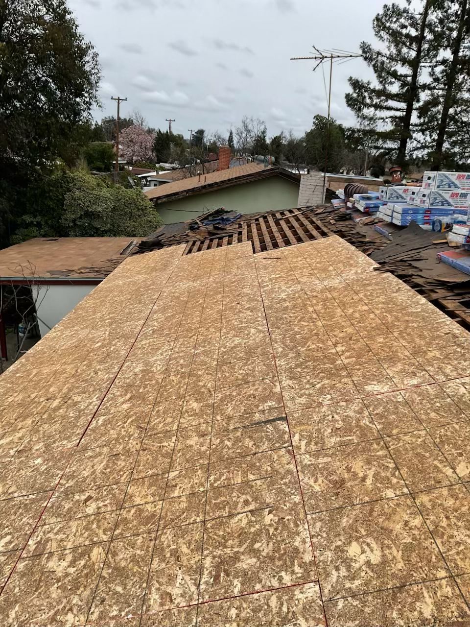 Roof under construction with plywood sheathing and exposed rafters, surrounded by trees and neighboring houses.
