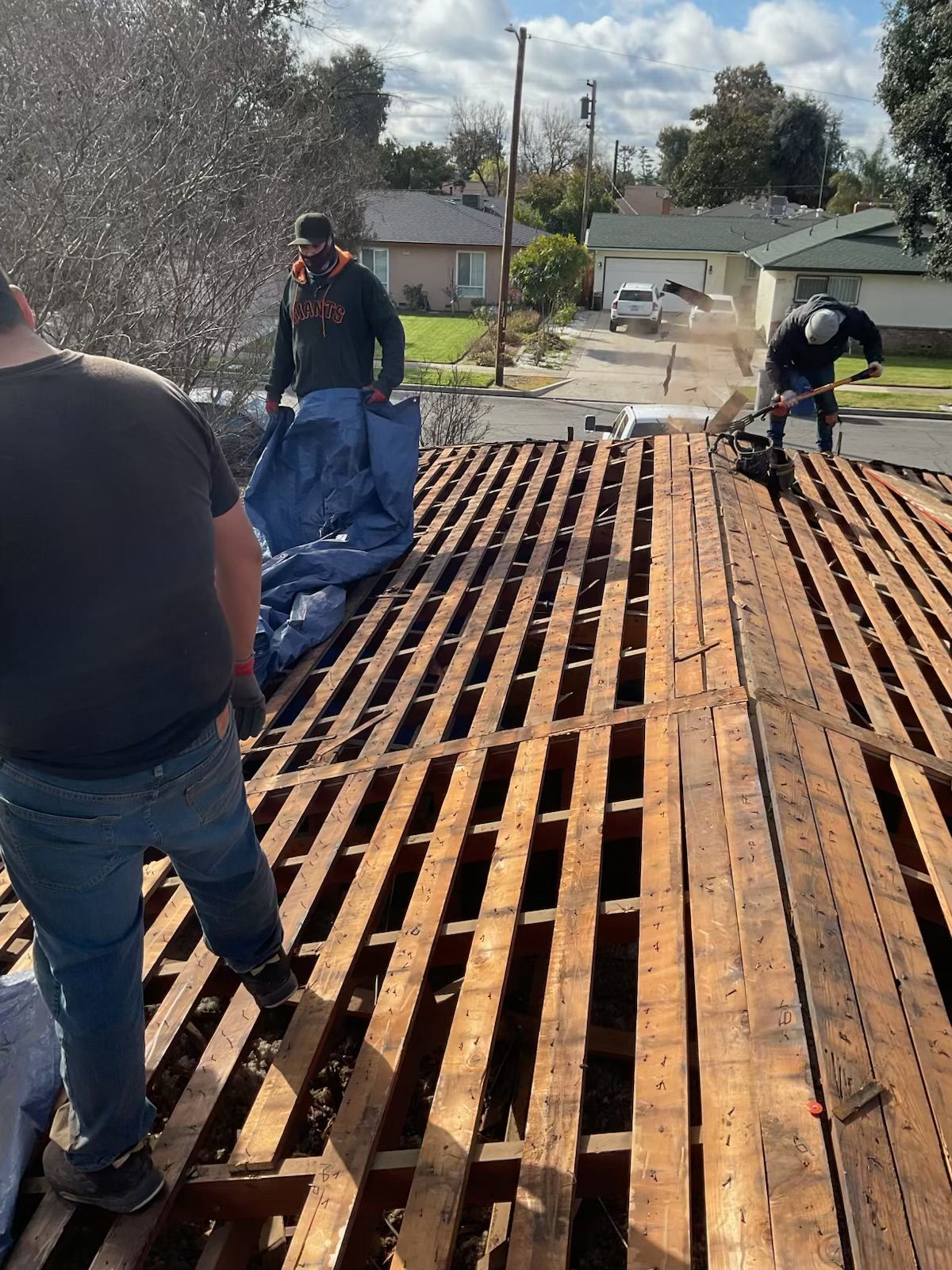 Workers replacing roof decking on a house, with exposed wooden rafters and debris on a sunny day.