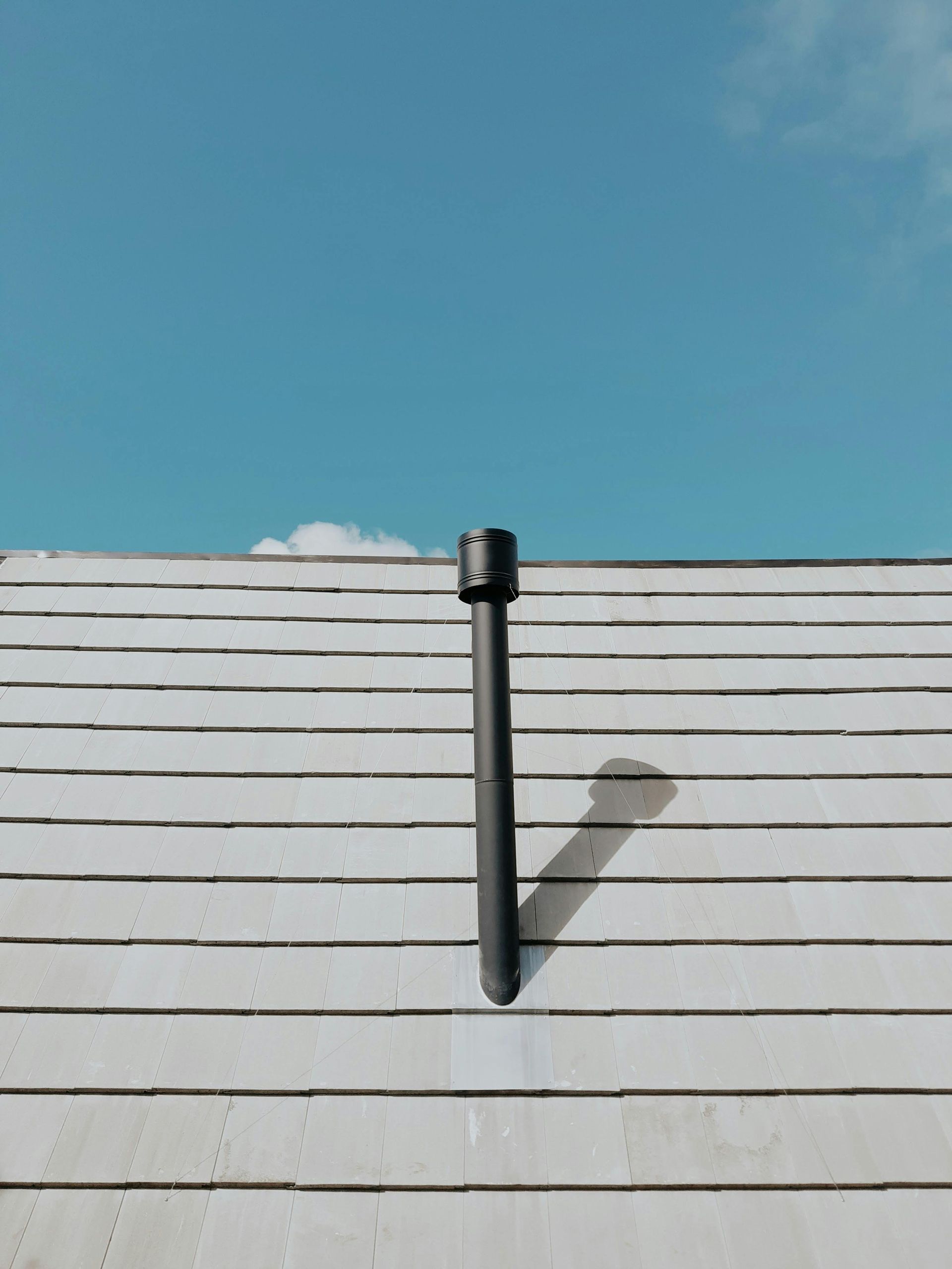 Black roof vent pipe on a white shingled roof under a clear blue sky.