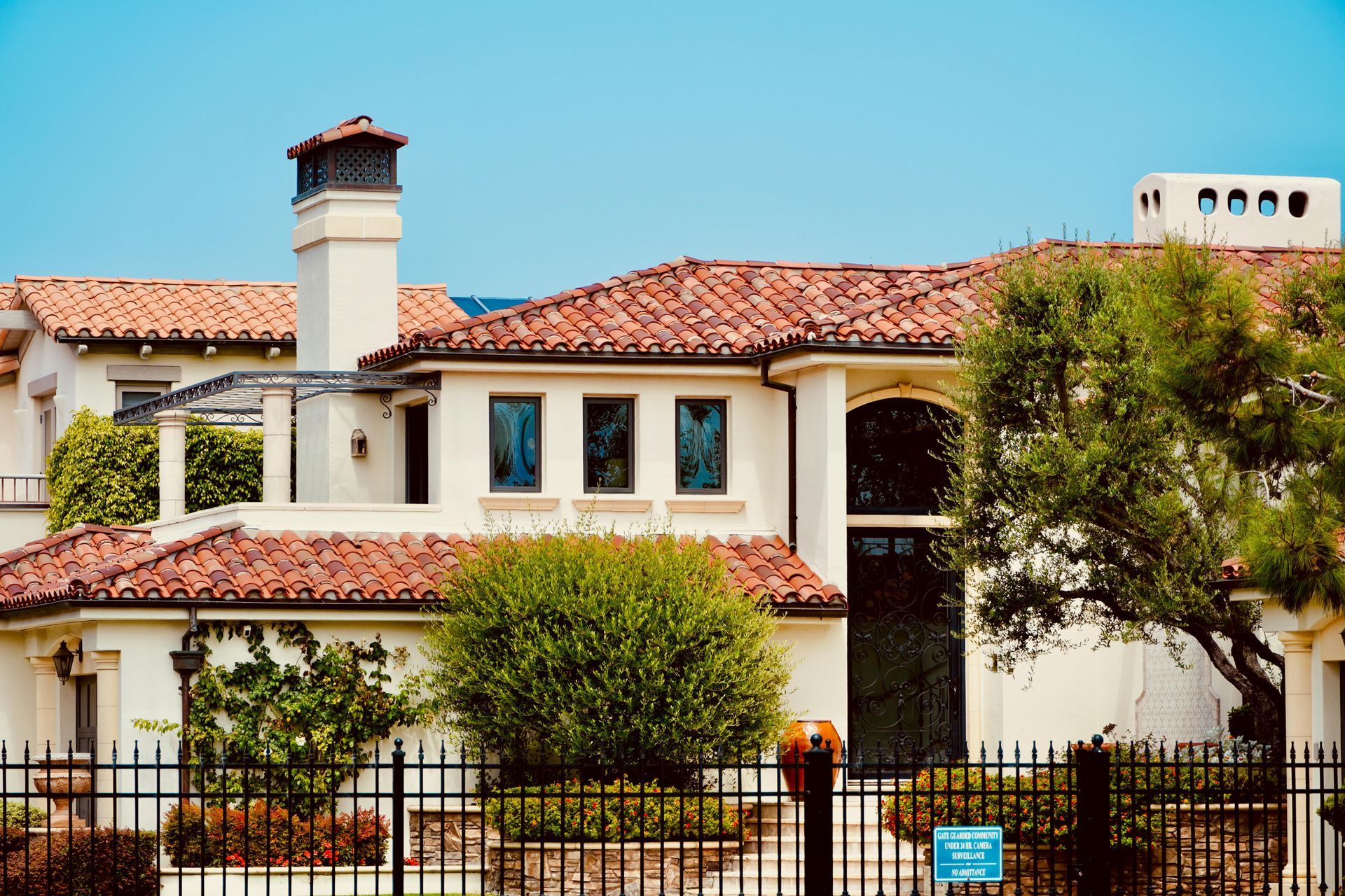 Two-story white stucco house with red tile roof, black fence, and leafy trees in front