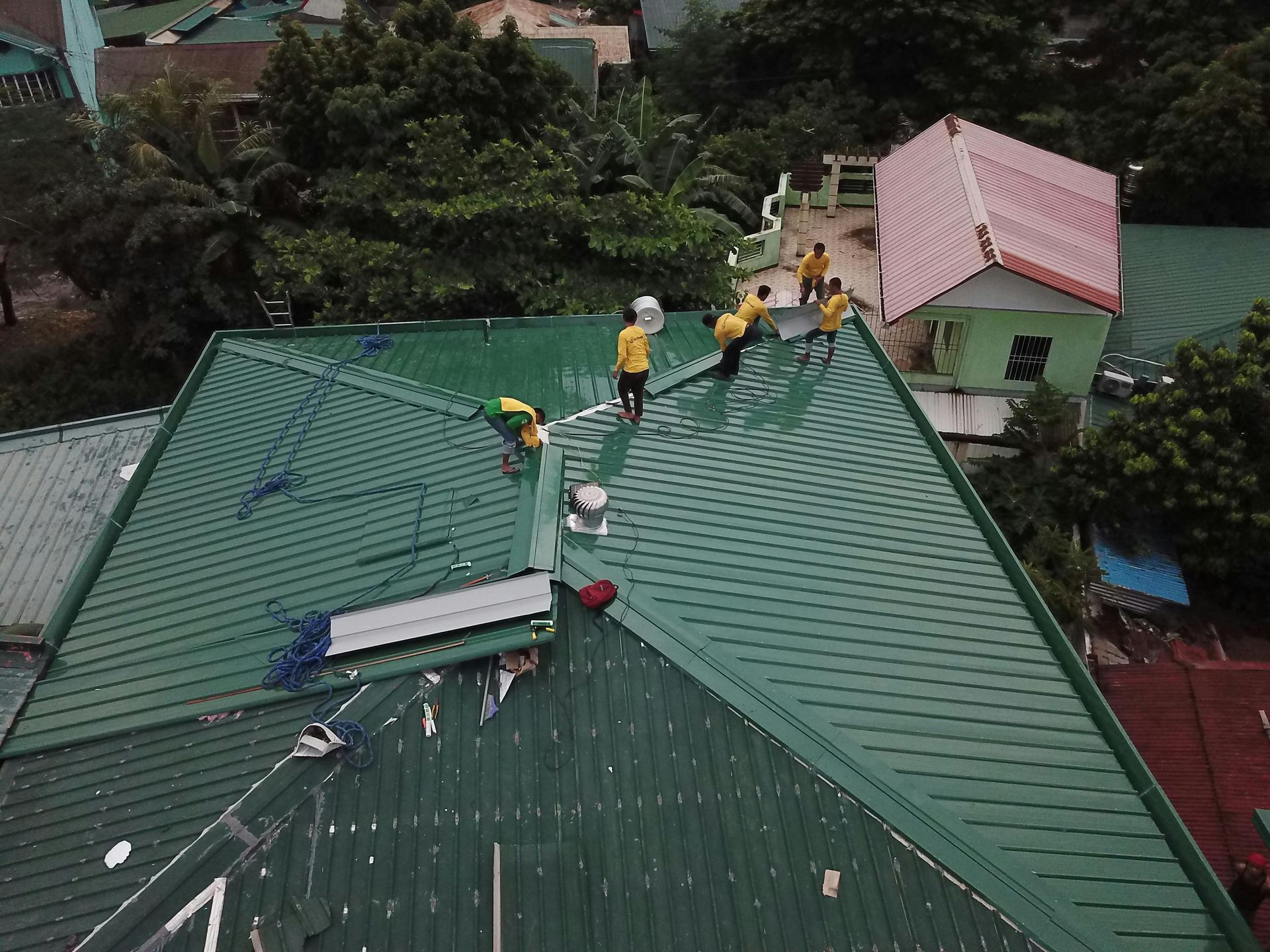 Workers on a green metal roof repairing panels, with a pink-roofed house nearby.