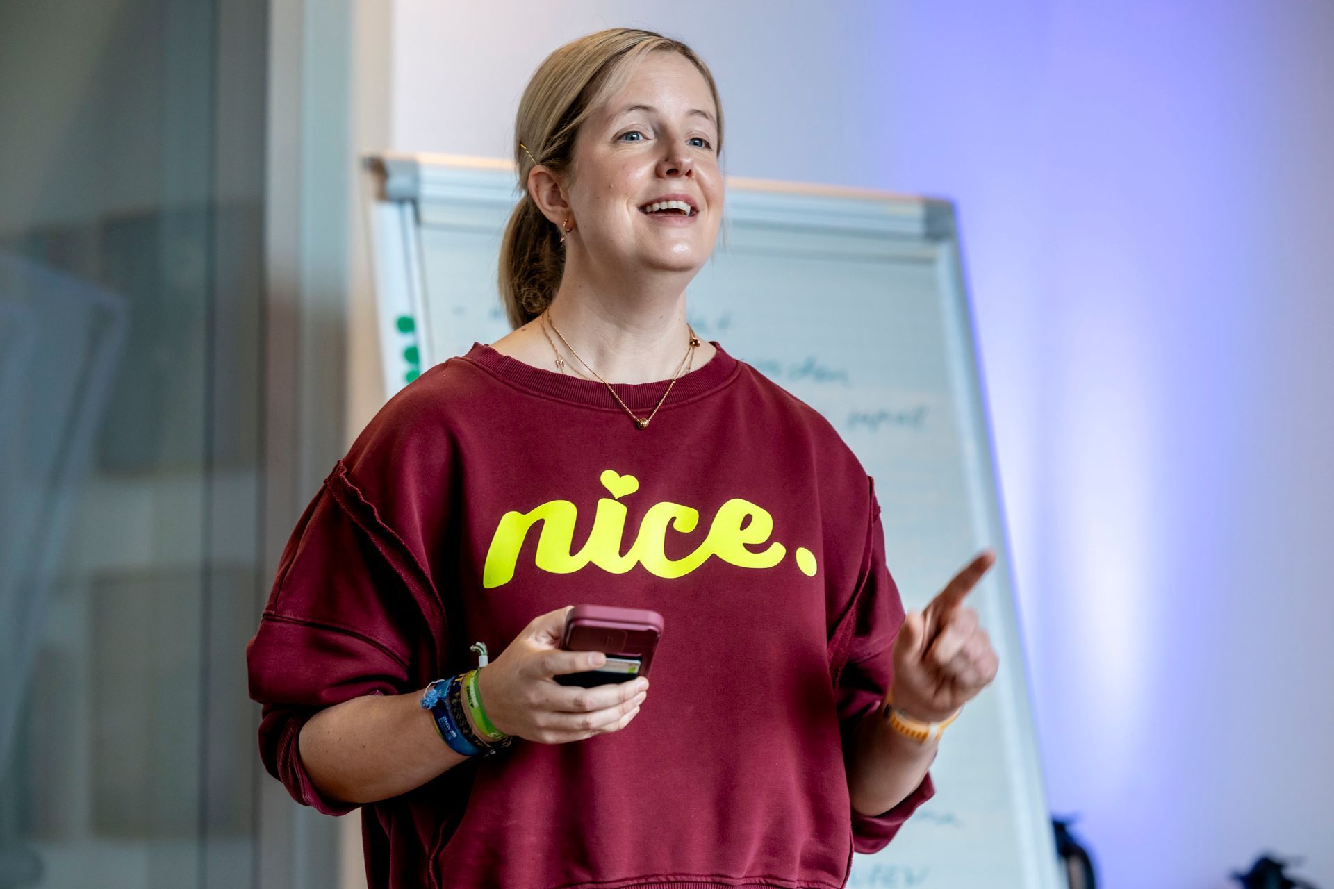 Frau in bordeauxrotem Sweatshirt mit der Aufschrift „nice“, die ein Handy hält und zeigt. Im Hintergrund eine Tafel.