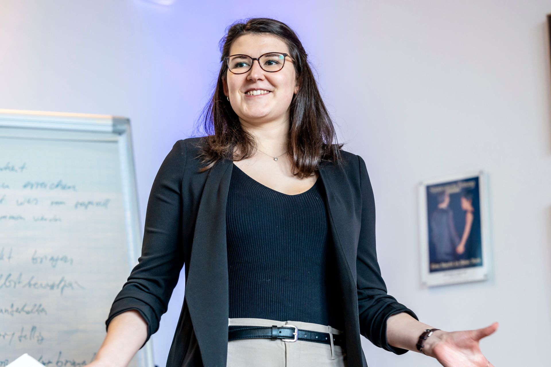 Frau mit Brille, schwarzem Blazer und dunklem Oberteil, lächelnd mit erhobenen Händen, in der Nähe einer Tafel.
