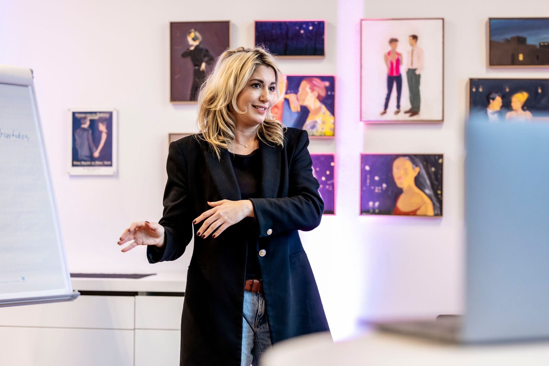 Eine Frau in einem schwarzen Blazer gestikuliert auf eine Tafel und scheint dabei einen Laptop anzusprechen; an der Wand hängt Kunst.