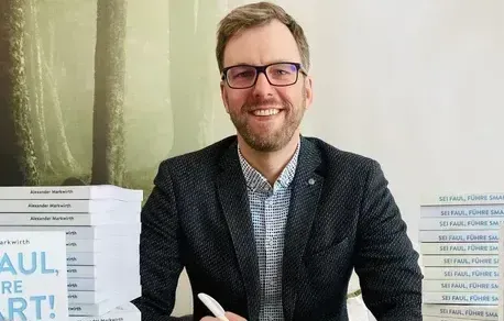 Ein Mann im Anzug und mit Brille sitzt vor einem Stapel Bücher.