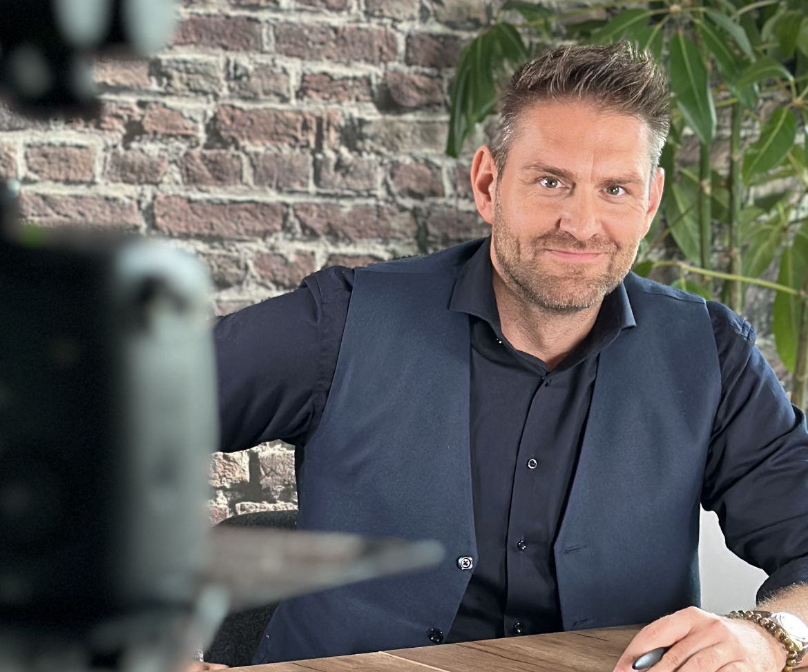 Mann am Schreibtisch mit Laptop und Telefon, klatscht in die Hände und lächelt in einem hellen Büro.