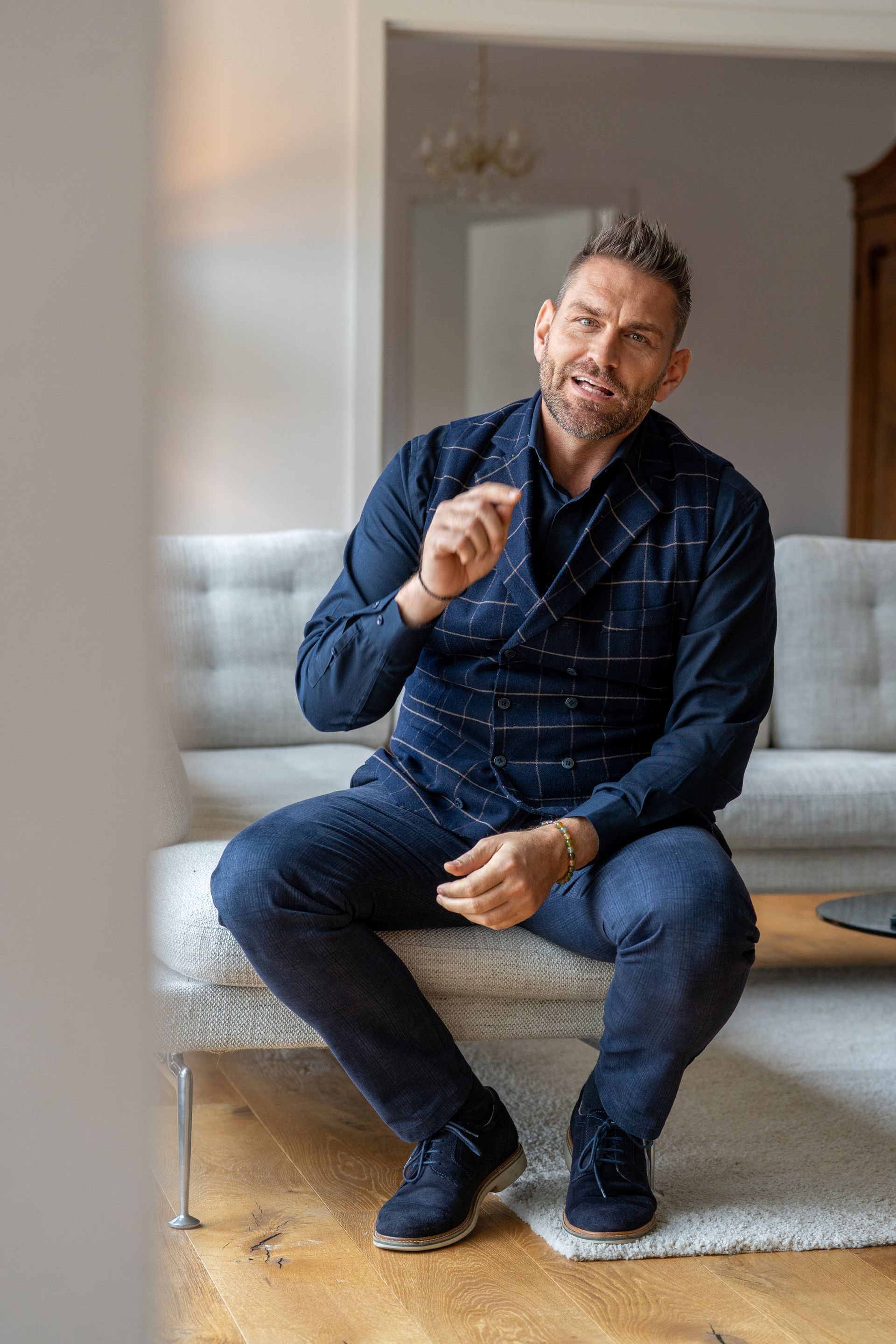 Mann klatscht in die Hände, lächelt in die Kamera, sitzt mit Laptop und Telefon an einem Tisch.