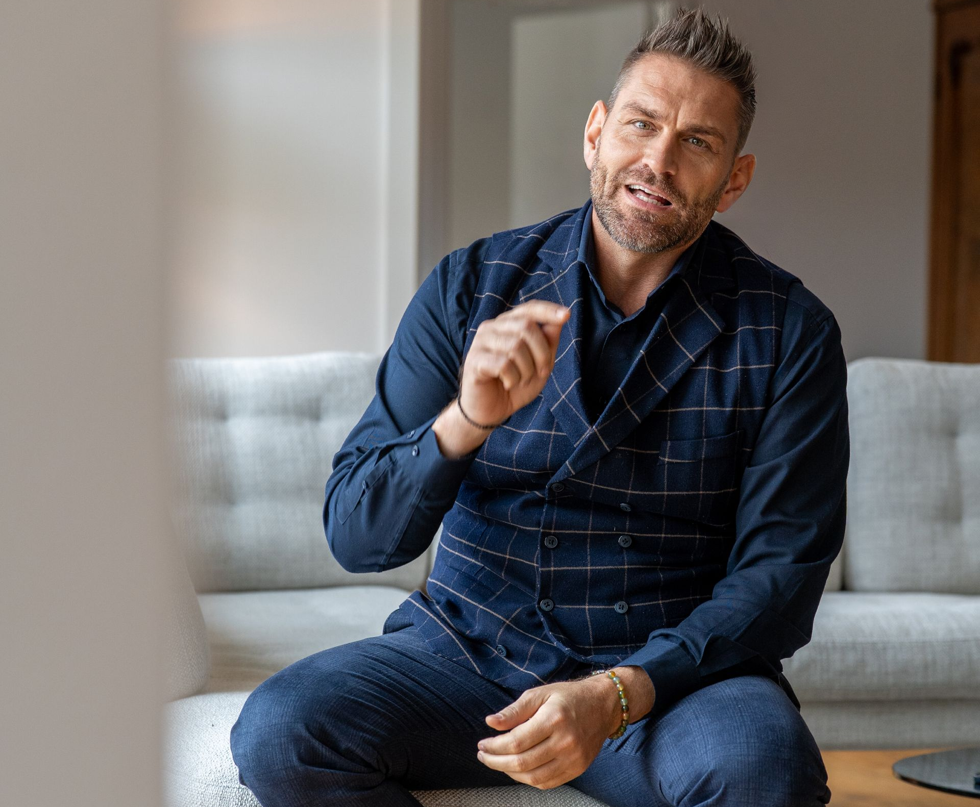 Mann im blauen Anzug gestikuliert, sitzt auf einem Sofa in einem Innenraum.