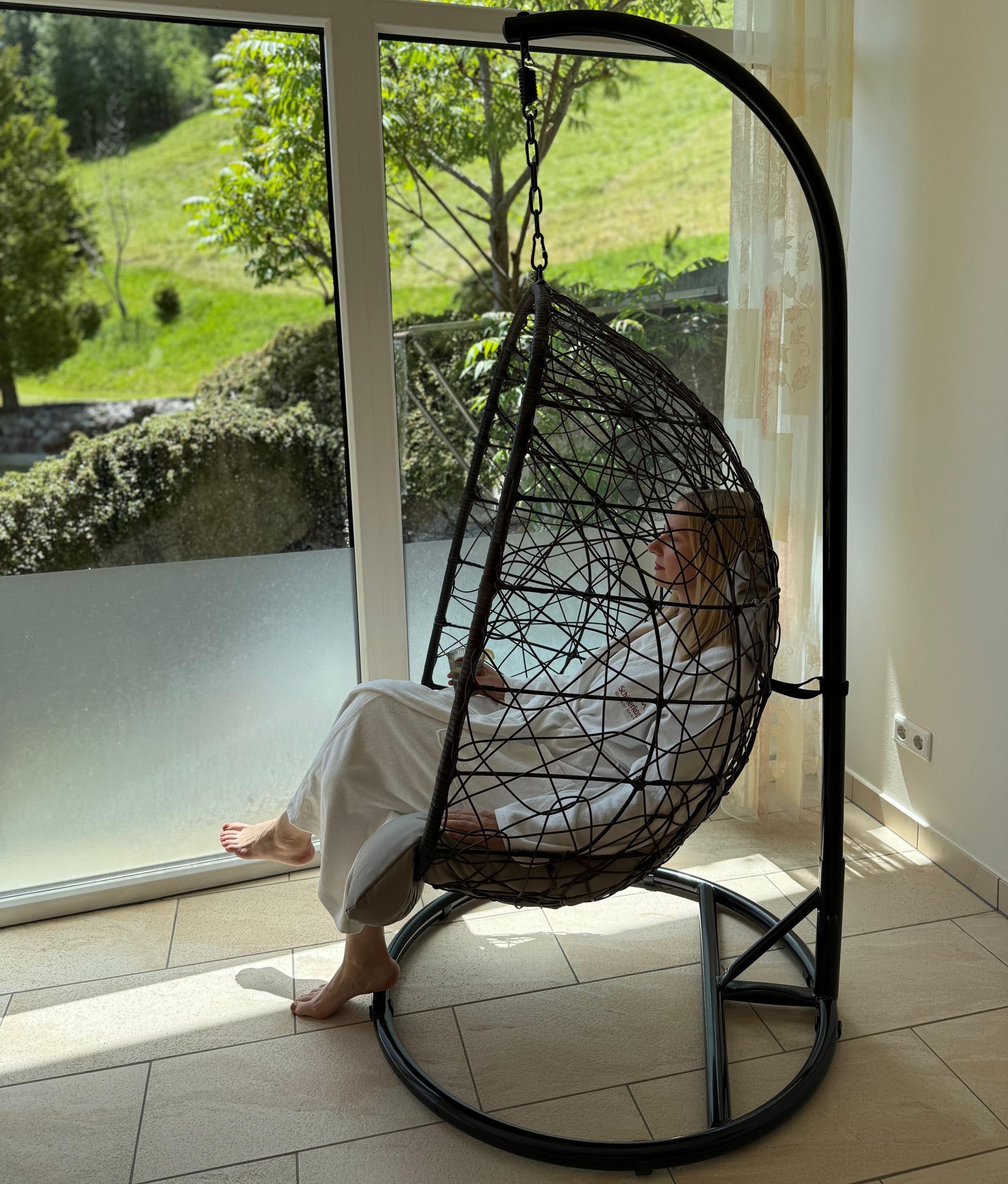 Eine Person sitzt in einem schwarzen Hängesessel am Fenster und blickt auf einen grünen Hügel.