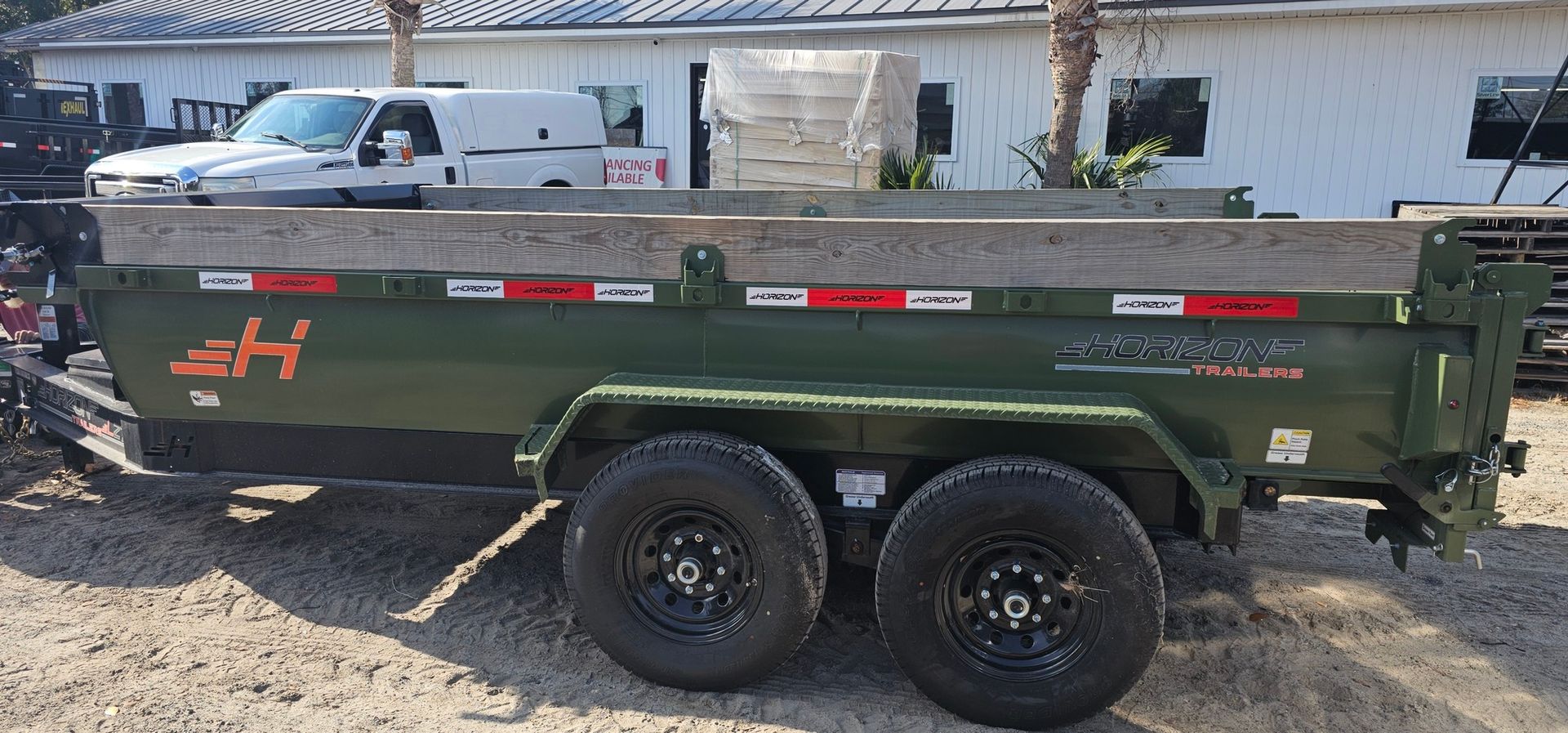 A dark green dual-axle dump trailer with wooden side extensions parked on a dirt lot in front of a white building.