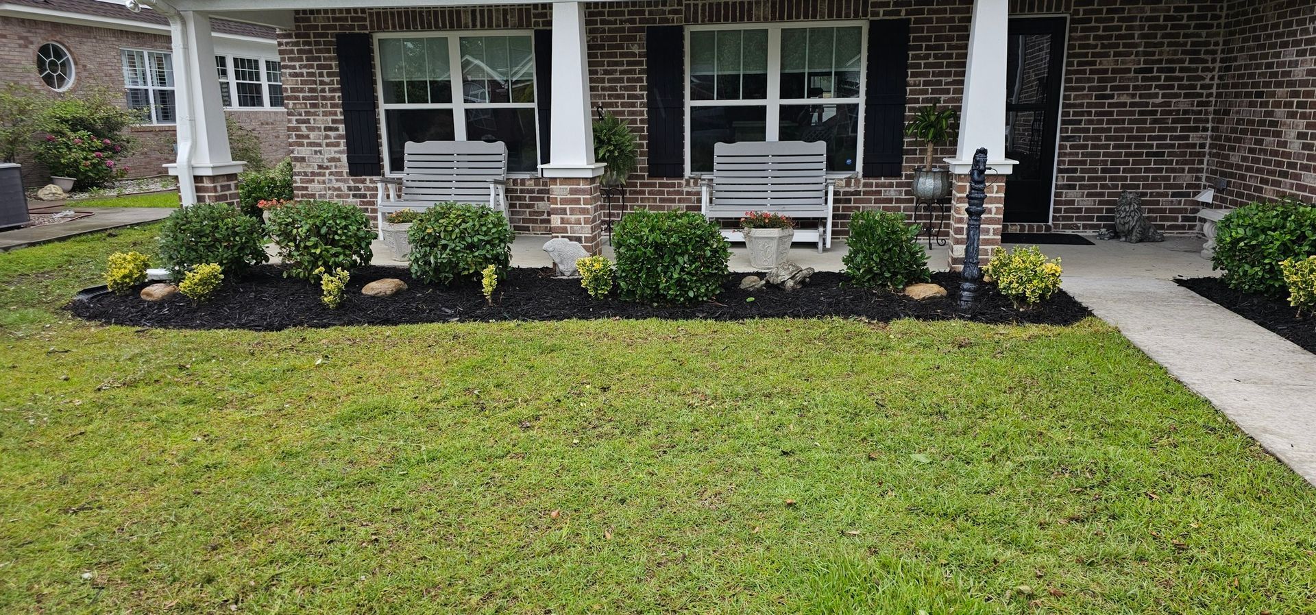 A brick house exterior features a covered front porch with two grey benches, neatly trimmed bushes, and black mulch.