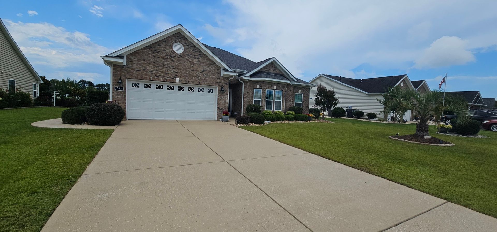 A single-story brick house with a two-car garage, concrete driveway, and landscaped front yard under a blue sky.