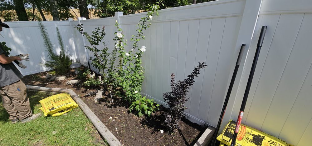 A person planting flowers and shrubs in a garden bed against a white vinyl fence.