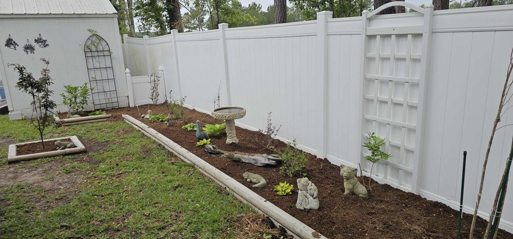 A backyard garden featuring a white vinyl fence, decorative trellises, a birdbath, and small plants in mulched beds.