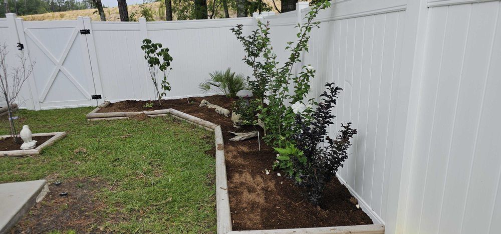 A white vinyl fence borders a backyard garden bed containing several small trees and shrubs with fresh mulch.