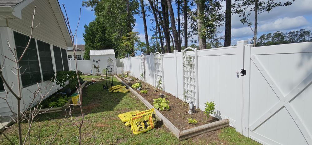 A side yard with a white vinyl fence, garden bed, trellis panels, and yellow bags of mulch near the house exterior.