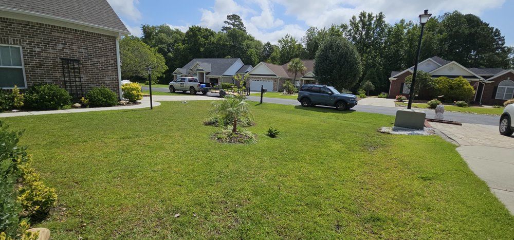 A suburban residential street on a sunny day with green lawns, brick homes, and parked cars under a blue, cloudy sky.