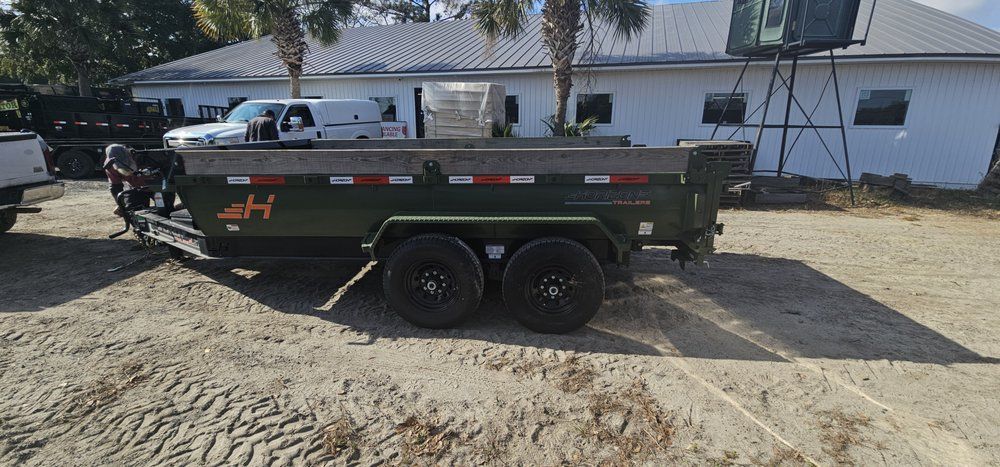 An olive green dump trailer with black wheels parked on a dirt lot in front of a white building.