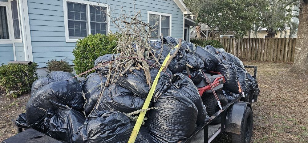 A utility trailer piled high with black garbage bags and debris, parked in front of a blue house.