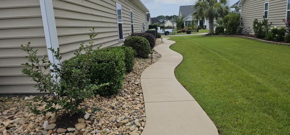 A curved concrete walkway leads along the side of a beige house with a rock garden and a neat green lawn.