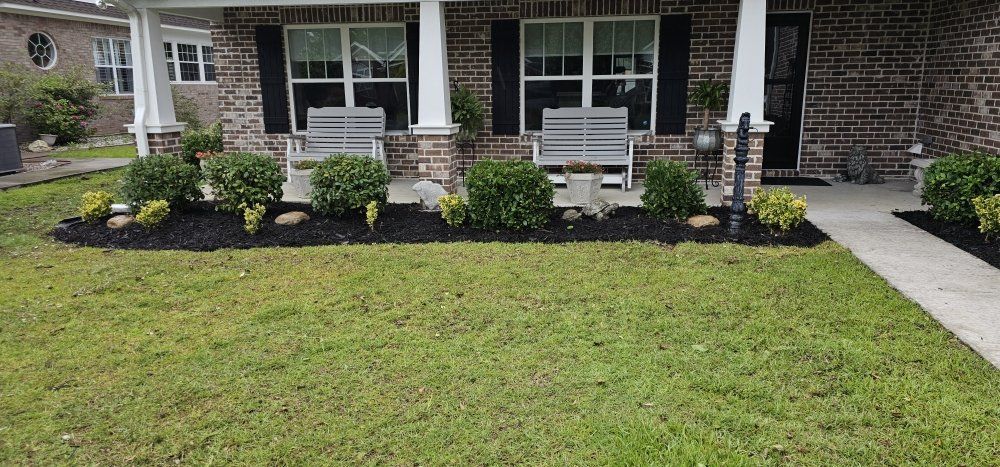 A brick house porch features two gray benches, dark shutters, and a front garden bed with green shrubs and black mulch.