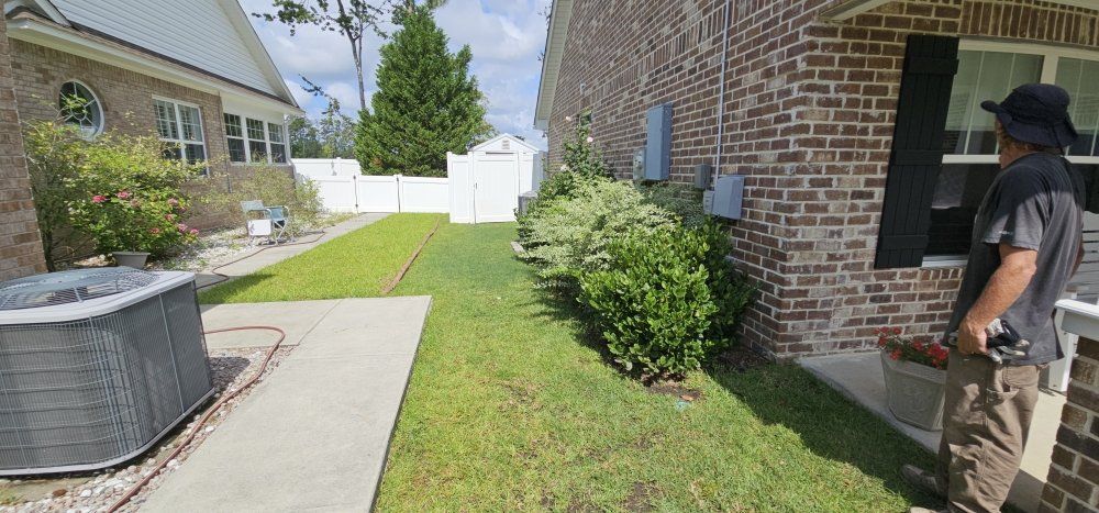 A person stands near a brick house next to a narrow grassy side yard with a concrete walkway and a central HVAC unit.