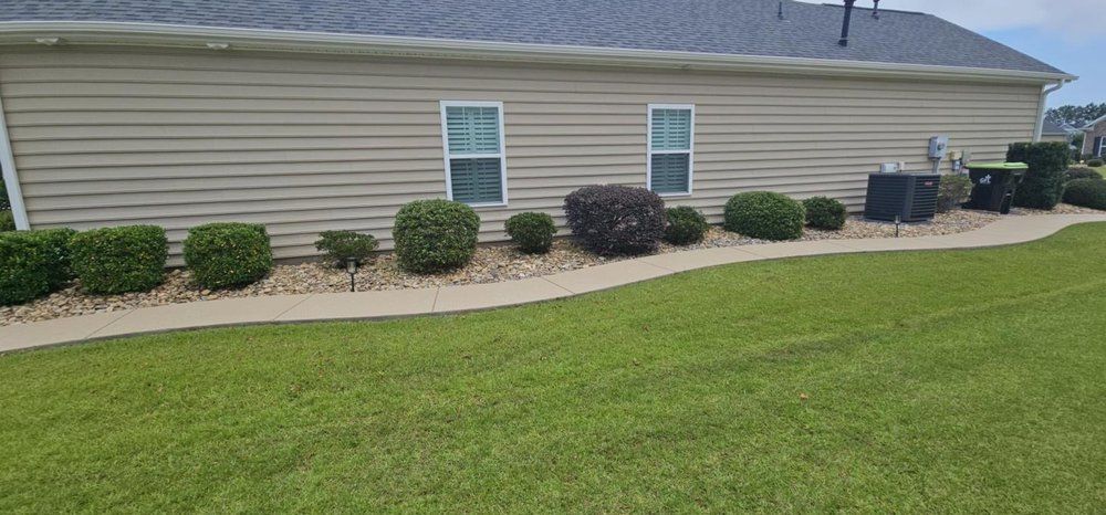 A beige, single-story house exterior with tan siding, two windows, and a landscaped garden bed along a concrete walkway.