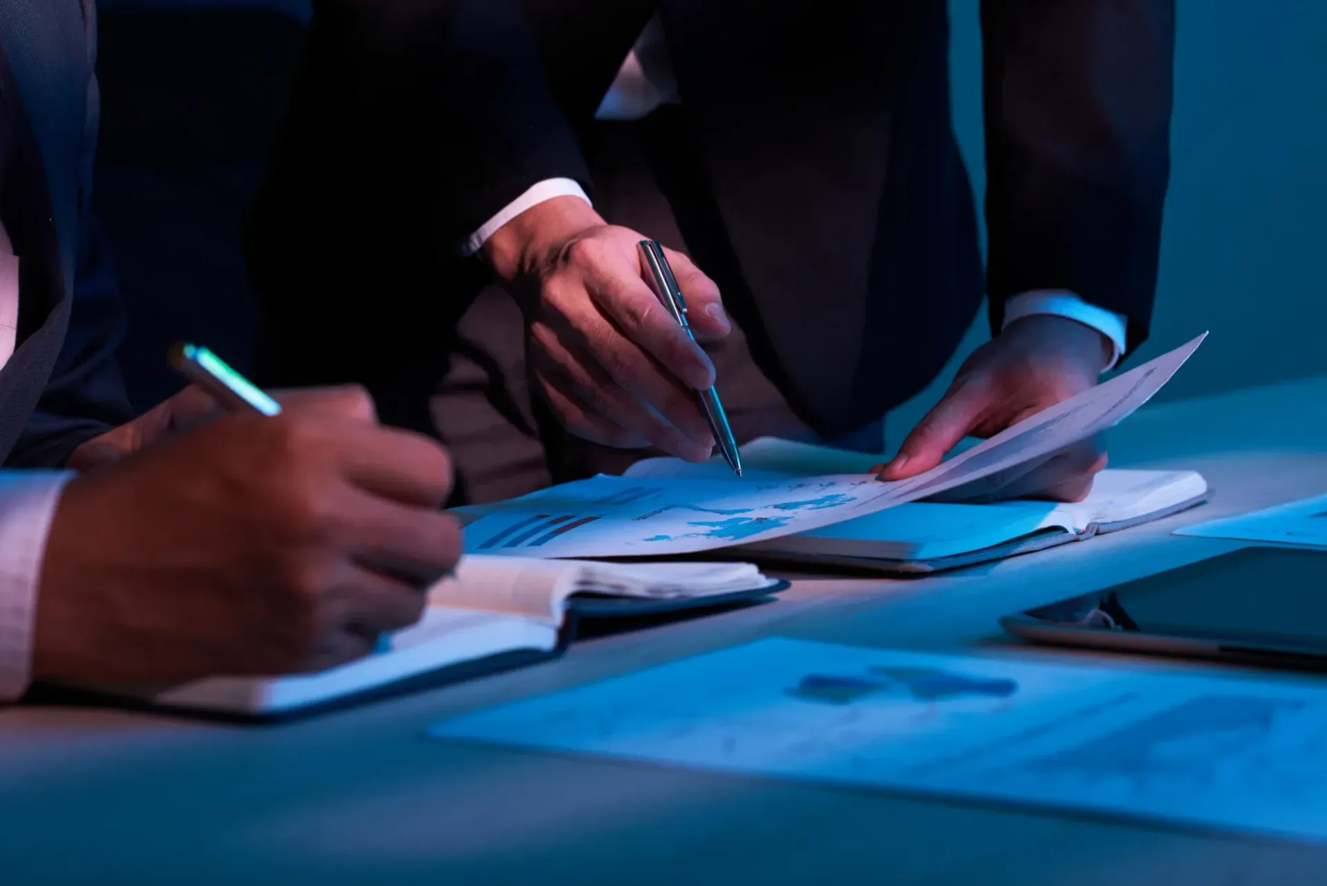 People in suits review documents, using pens, at a desk.