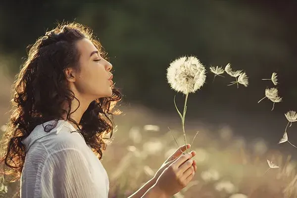 A woman is blowing dandelions in a field.