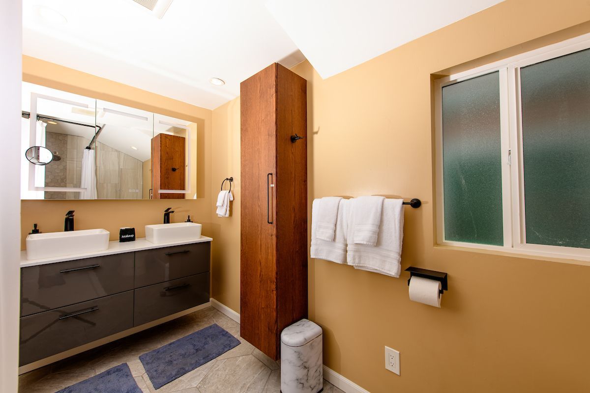 A bathroom with two sinks , a trash can , a mirror and a window.