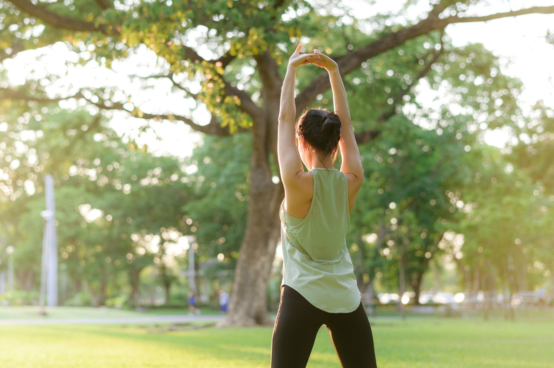Woman stretching arms up in a sunny park with a large tree in the background.