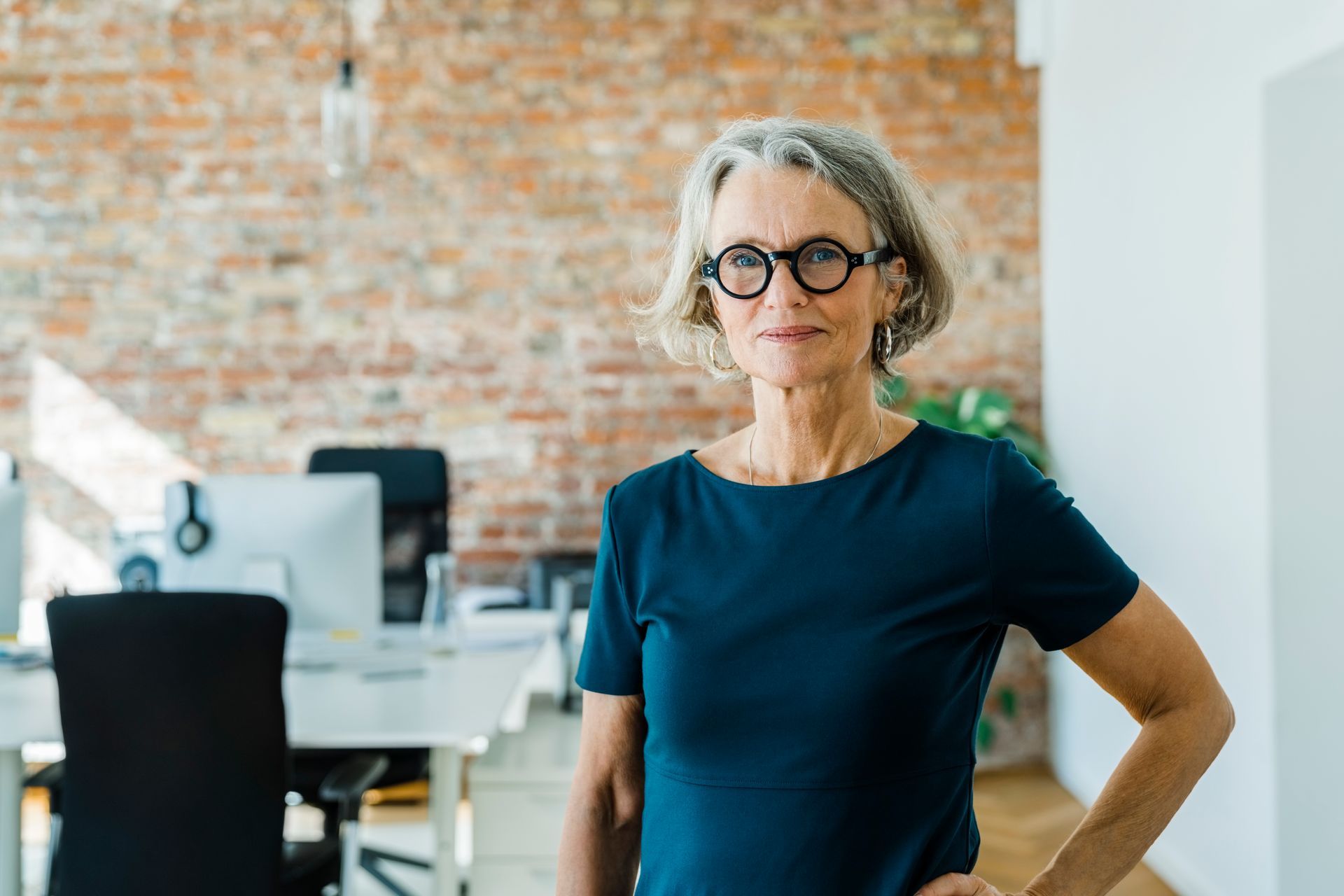 A woman wearing glasses is standing in an office with her hands on her hips.