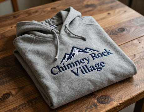 Embroidered hoodie with Chimney Rock Village branding folded on a rustic wooden table.