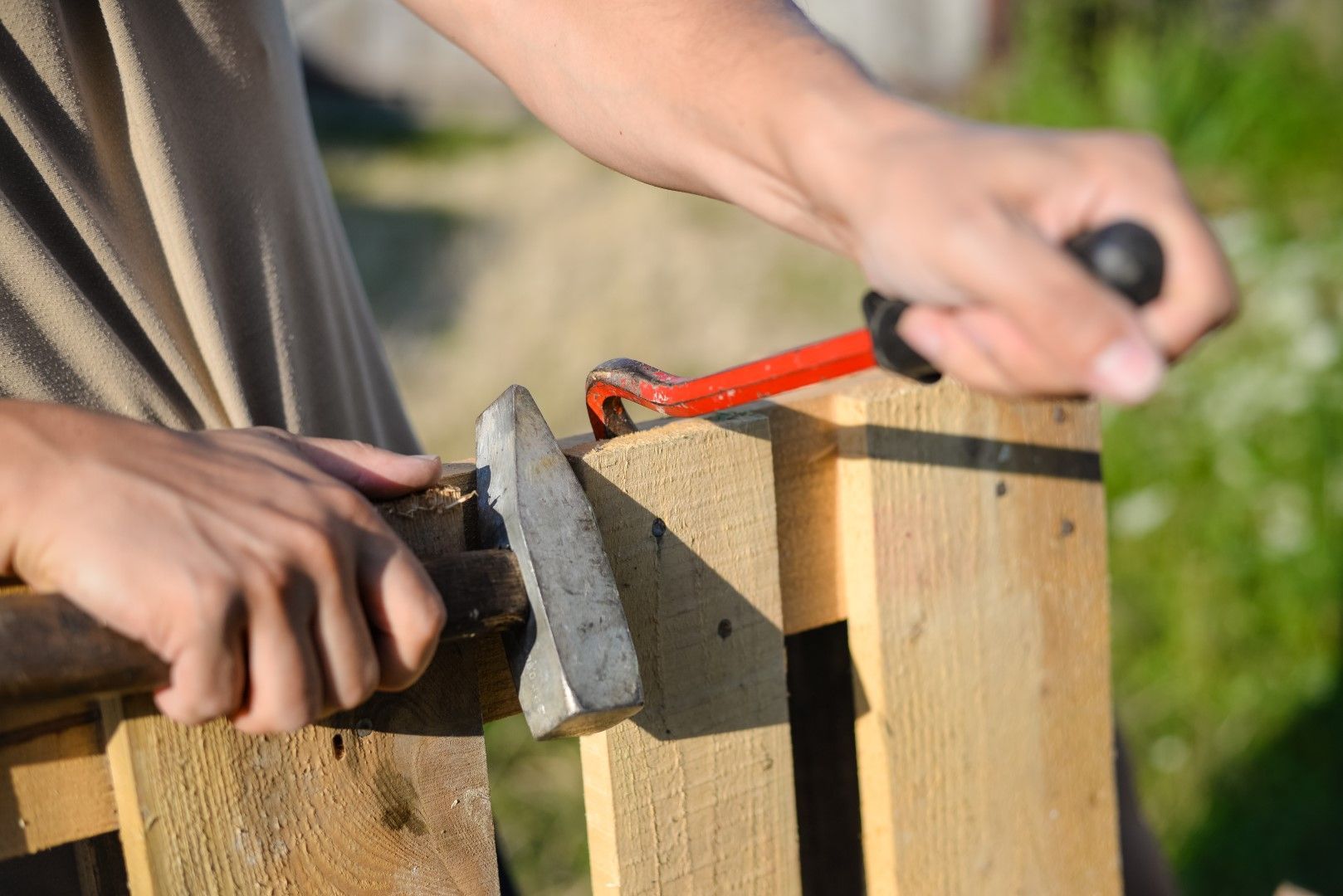 Person using a pry bar to remove boards from a wooden pallet, outdoors.